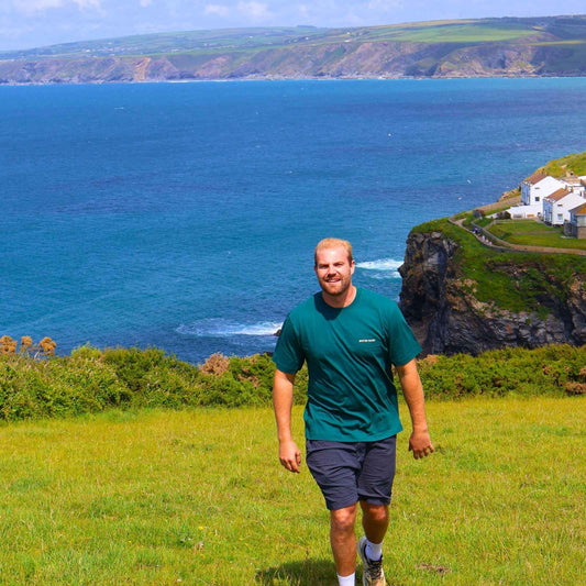 Man walking on a grassy field with a scenic ocean view and cliffs in the background.