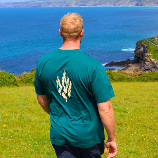 Man wearing a green t-shirt with a logo standing on a grassy field overlooking the ocean.