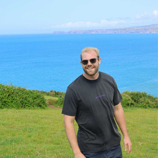 Man wearing sunglasses and a black t-shirt standing in a grassy field with a blue ocean and sky in the background.