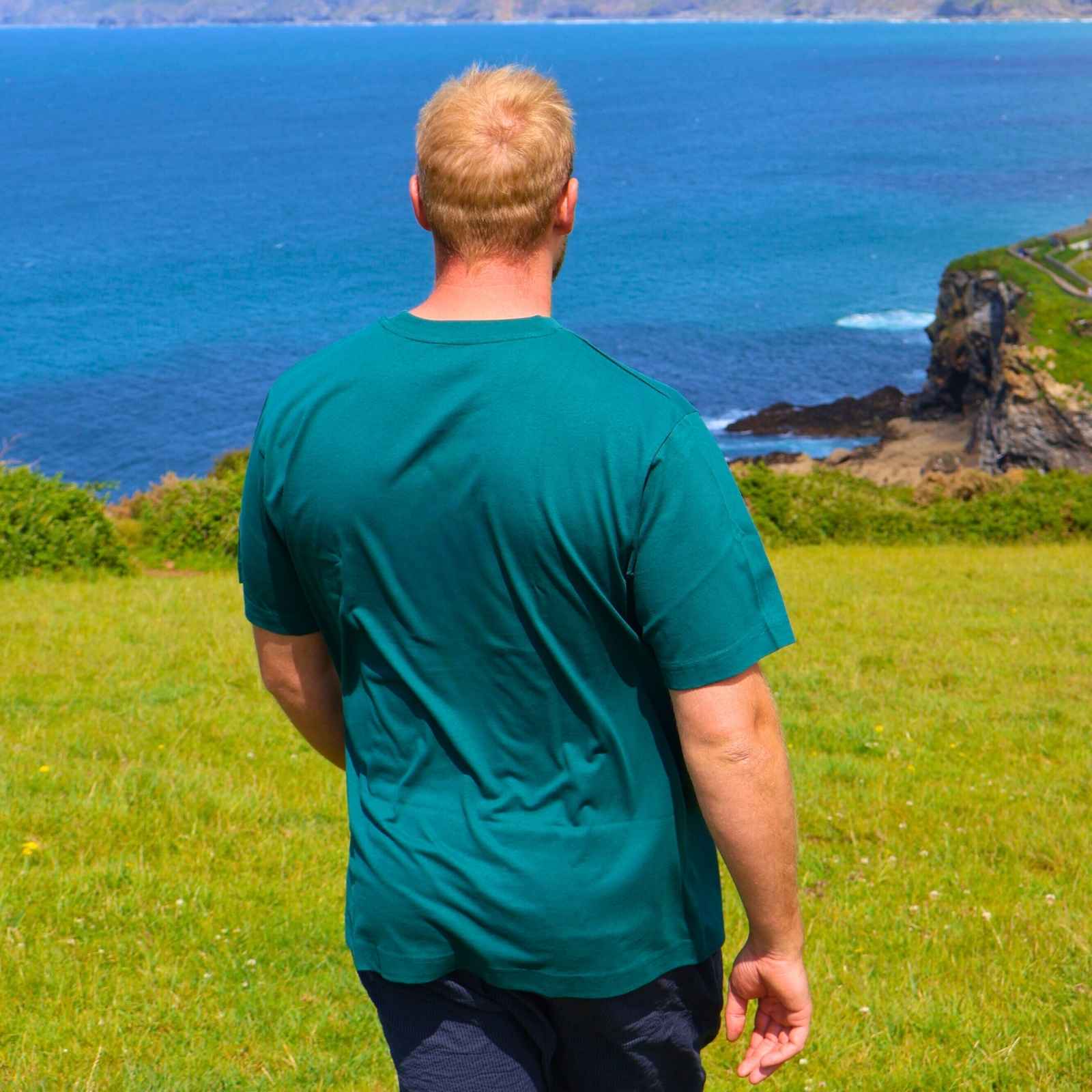 Man in a green shirt standing on a grassy field looking at the ocean.