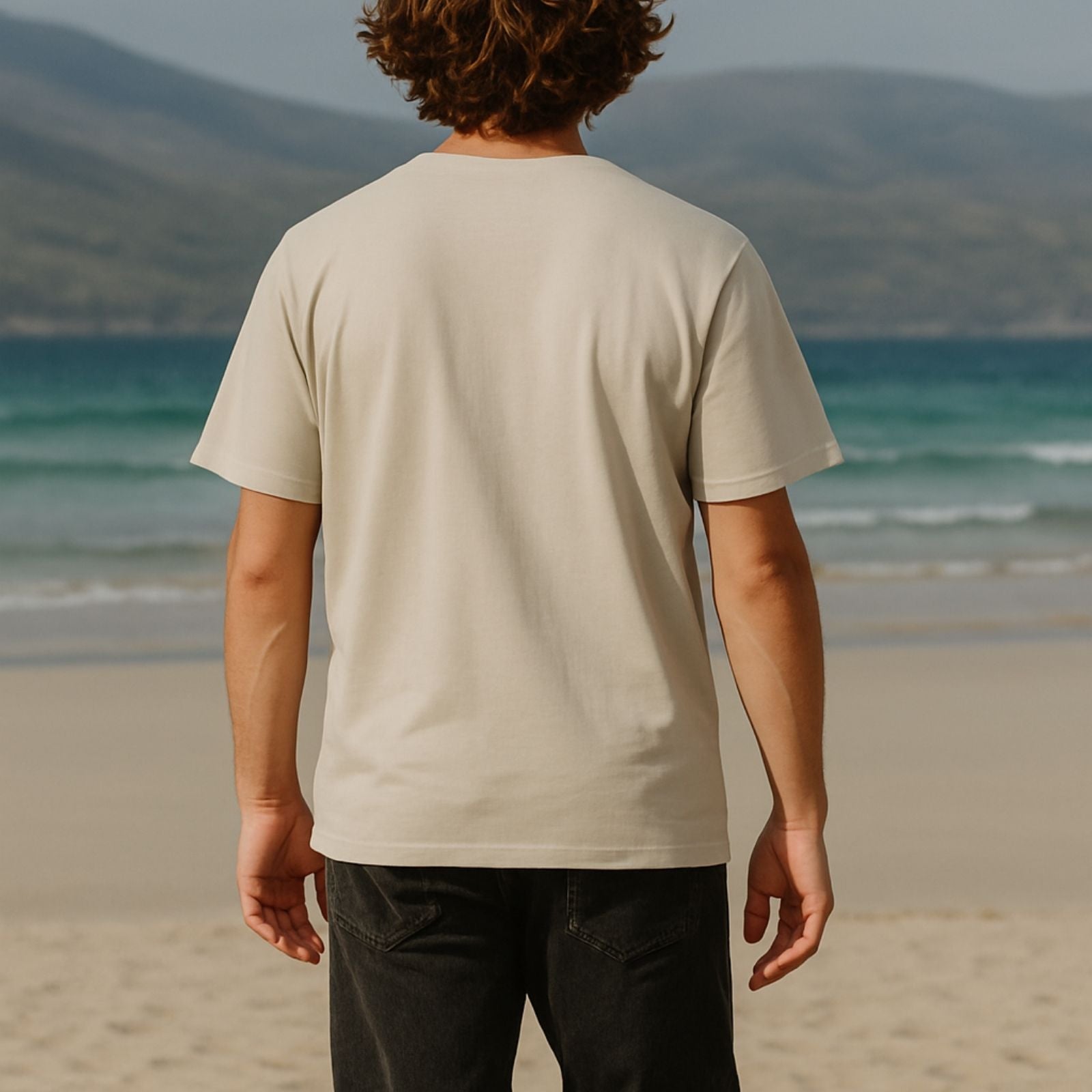 Person wearing a beige t-shirt on a beach with mountains in the background