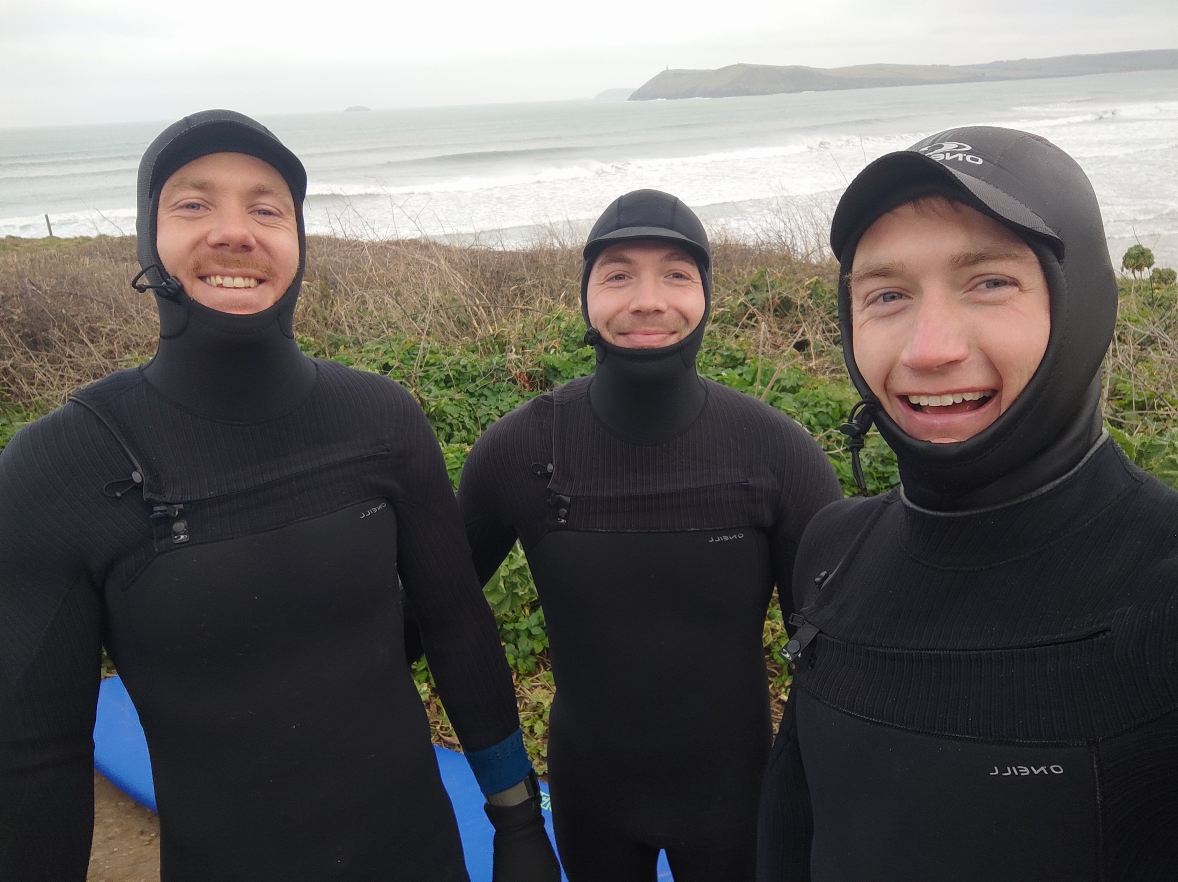 Three people in black wetsuits standing outdoors with a body of water in the background.